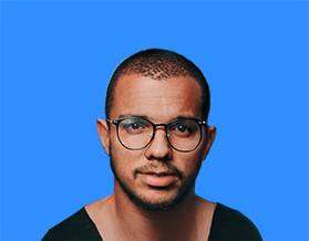 A man with light stubble and closely cropped hair wearing black-framed glasses, against a solid blue background. he looks directly at the camera with a serious expression.