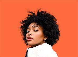 A woman with curly black hair and earrings looks over her shoulder against a bright orange background. she wears a white blouse and has a calm expression.