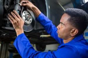 A mechanic in a blue uniform inspects and repairs the brake system on a vehicle suspended on a lift in an auto repair shop.