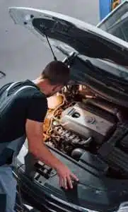 A man wearing a backpack inspects the engine of a car, which is open in a garage setting. his focus is on the vehicle's internal components.