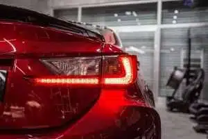 Close-up of a red car's illuminated taillight, with a glossy finish reflecting a dimly lit garage. a hint of a treadmill is visible in the background on the right.