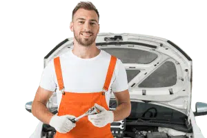A smiling male mechanic in an orange jumpsuit and white gloves holding a wrench, standing in front of a car with its hood open.